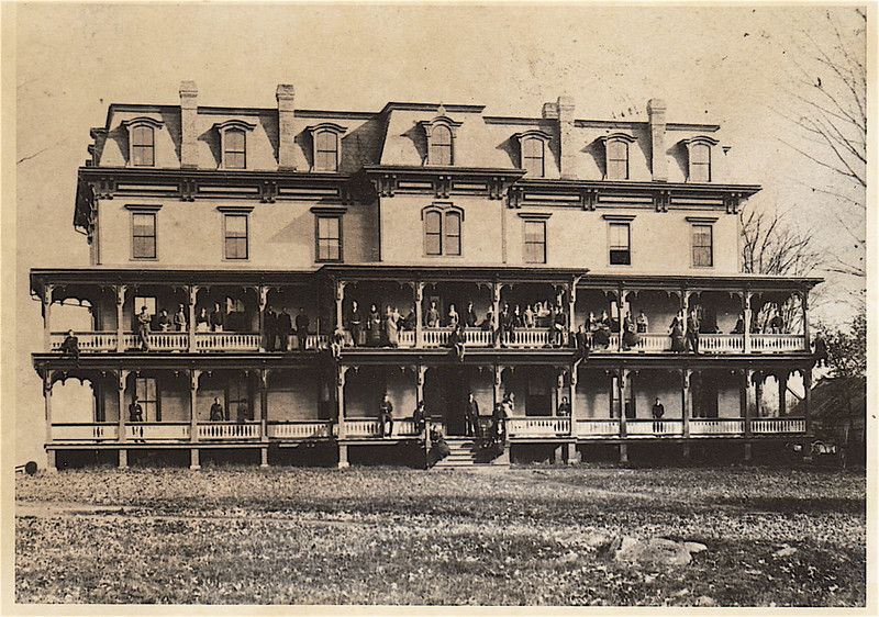a old wooden hotel building with two levels of wrap-around porches and people standing on both sets of porches