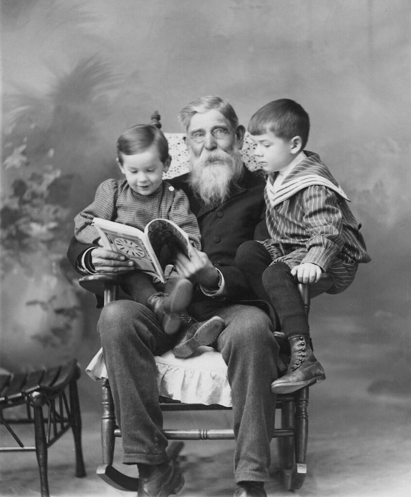 A black and white photo of an elderly man with a gray beard sitting in a chair and holding a book open. He is looking at the camera while the two children on his lap are looking at the book..